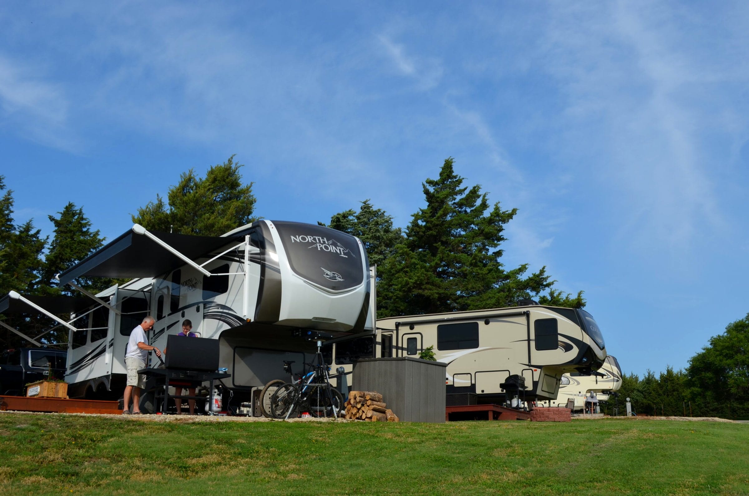 A large fifth-wheel camper set up on a grassy site under evergreen trees